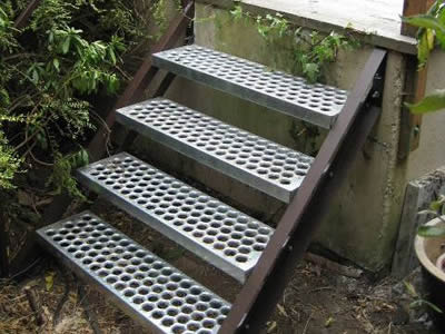 4 safety grating plates form a stair under a platform; surroundings on one side of the stair include many plants.
